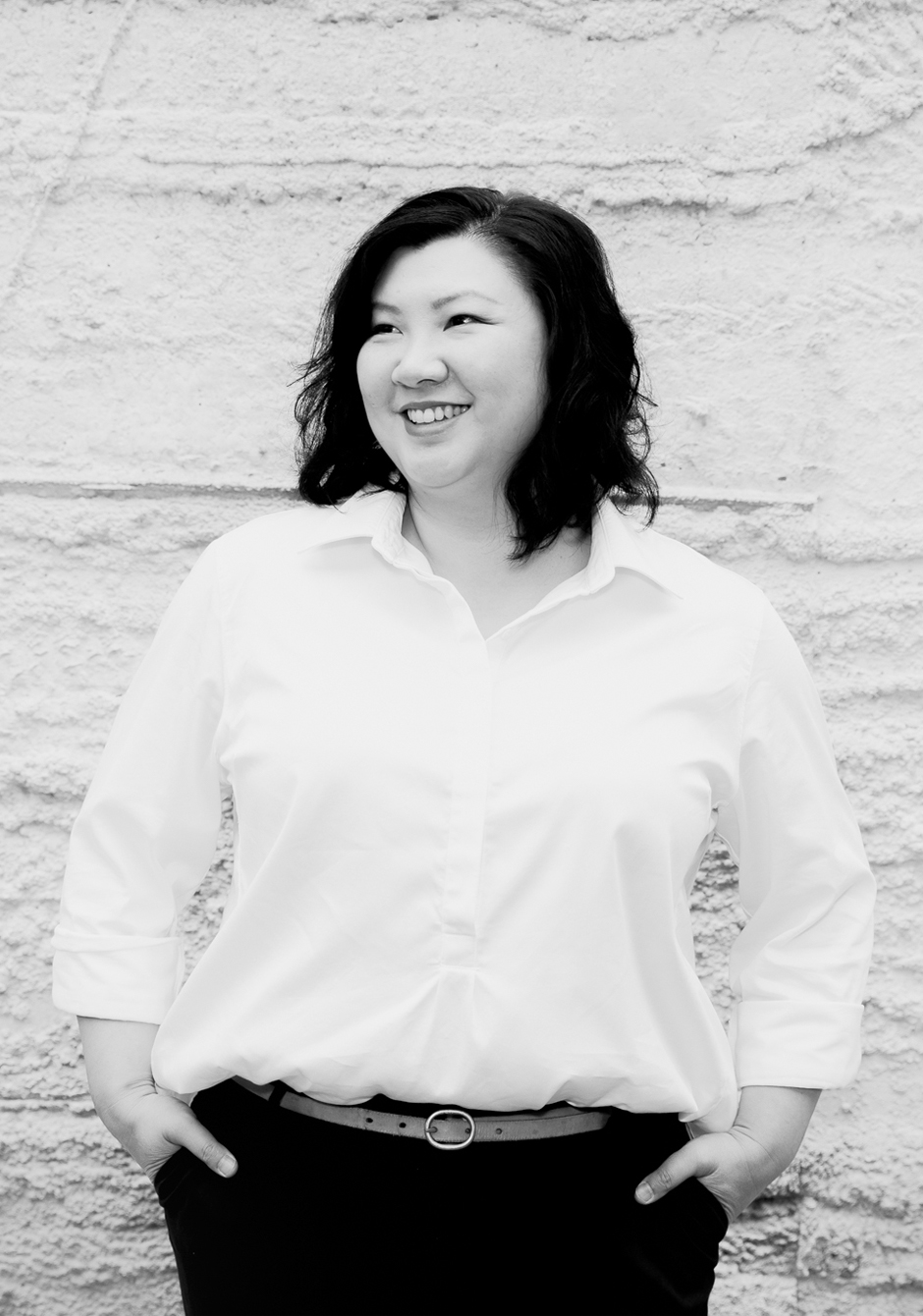 A woman stands facing the camera looking away. She has her hands in her pant pockets and she is smiling.