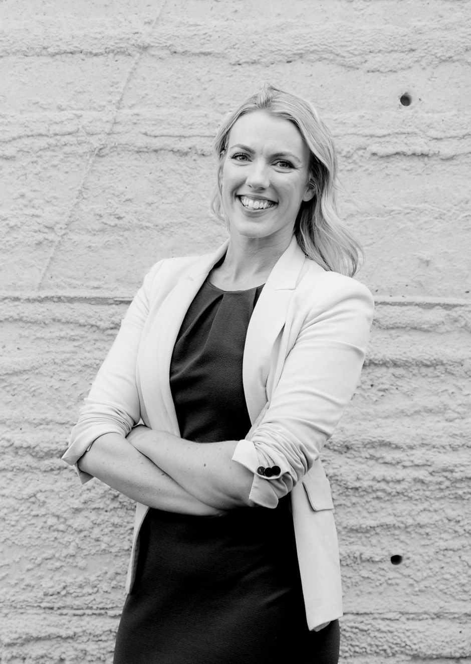 A black and white image of a woman. She is standing facing the camera and smiling with her arms crossed.